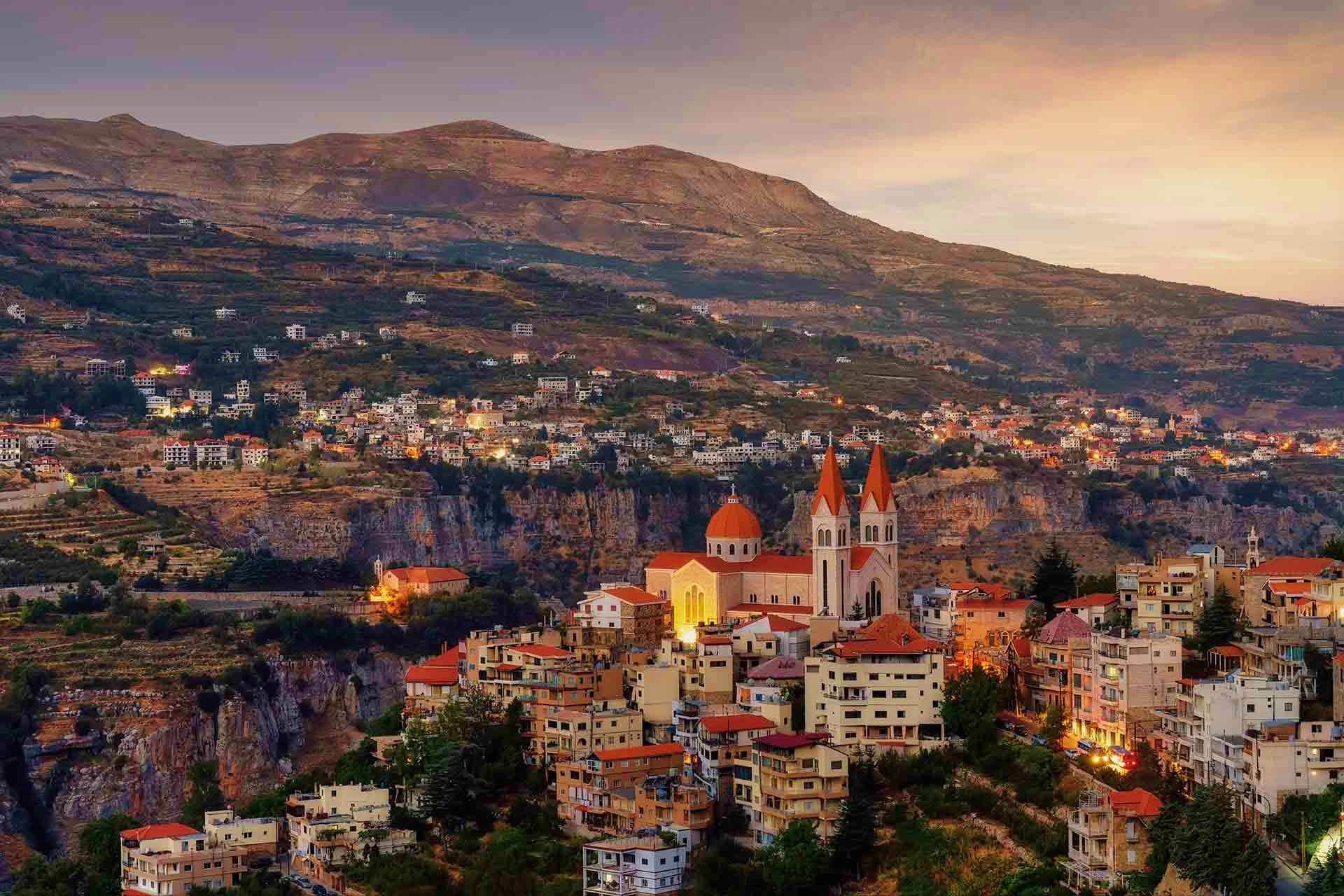 Aerial view of Harissa, Lebanon at dusk with illuminated white church with red-orange domes overlooking coastal town nestled